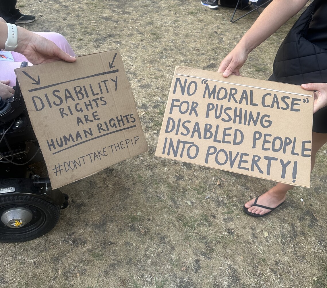 two cardboard protest signs that read - &#39;Disability rights are human rights #DontTakeThePIP&#39; and &#39;No moral case for pushing Disabled people into poverty!&#39;.
